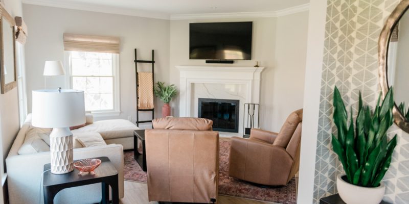 A warm, welcoming living room featuring a white marble fireplace, wall-mounted TV, leather recliner, beige sofa, and layered rugs — designed for comfort, style, and seamless daily living in this remodeled home.