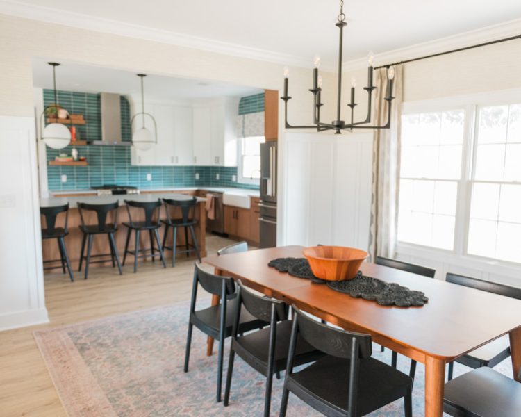 A bright, airy dining zone featuring a rich wood table, black upholstered chairs, and a statement chandelier — framed by a clear view into the adjacent kitchen with its vibrant teal tile backsplash and floating shelves, emphasizing open-concept flow and style.