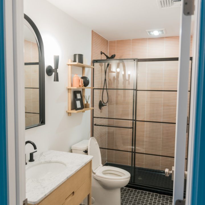 A stylish bathroom featuring a light wood vanity, white quartz countertop, matte black fixtures, geometric black-and-white floor tile, and a frameless glass shower with warm-toned tiled walls — blending modern function with bold pattern and texture.