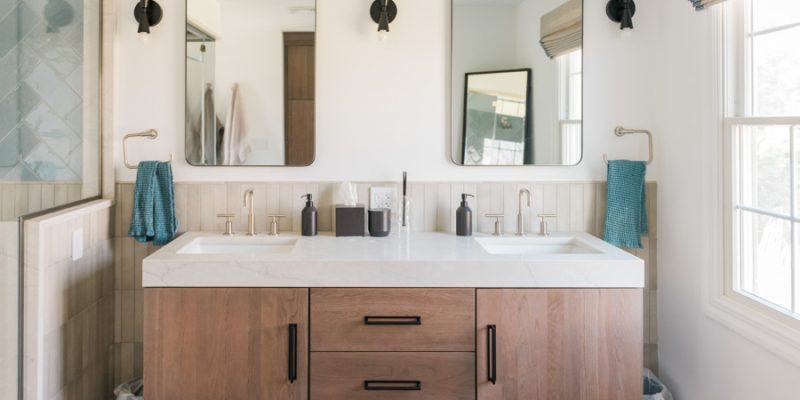 A sophisticated master bathroom featuring a floating wood-grain double vanity with white quartz countertop, matte black hardware, dual rectangular mirrors, and brass-finished faucets — blending warmth, contrast, and clean lines for a high-end, functional retreat.