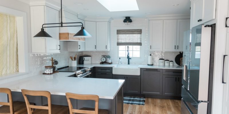 A panoramic shot of a fully remodeled kitchen featuring a large white countertop island, wood base, modern pendant lights, stainless steel appliances, and sightlines to the adjacent living area — emphasizing openness, connectivity, and high-impact style.